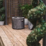 Dark rustic wooden gas fire pit placed on a wooden deck in an outdoor garden setting.