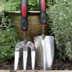 Gardening tools set, including a stainless steel hand trowel and hand fork, placed in a garden setting with lush greenery