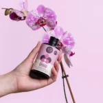 Hand holding Wild the Water Orchid Care nutrient solution bottle near a blooming orchid, formulated for orchid health.