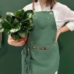 Person wearing We The Wild green gardening apron with pockets, holding a potted plant, ideal for organized and practical gardening.